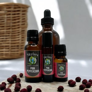 May include: Three brown glass bottles with black caps and labels that say "Skylara Essentials Pink Peppercorn". The bottles are arranged in a row on a wooden surface with pink peppercorns scattered around them.