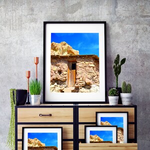 May include: A rustic stone building with a wooden door and a blue sky in the background. The building is framed in a black picture frame and is displayed on a wooden dresser with other framed pictures.