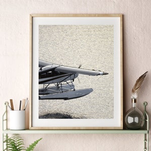 Könnte beinhalten: Ein gerahmtes Foto eines Wasserflugzeugs, das über einem Gewässer abhebt. Das Flugzeug ist weiß mit schwarzen Akzenten und hat zwei Schwimmer. Das Wasser ist hellblau und der Himmel ist hellblau. Das Foto hängt an einem Regal mit anderen Dekorationsartikeln.