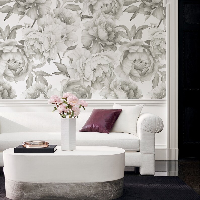 May include: A white couch with a burgundy throw pillow sits in front of a wall with a floral wallpaper pattern. The wallpaper features large, gray peonies with white centers. A vase with pink flowers sits on a white oval coffee table.