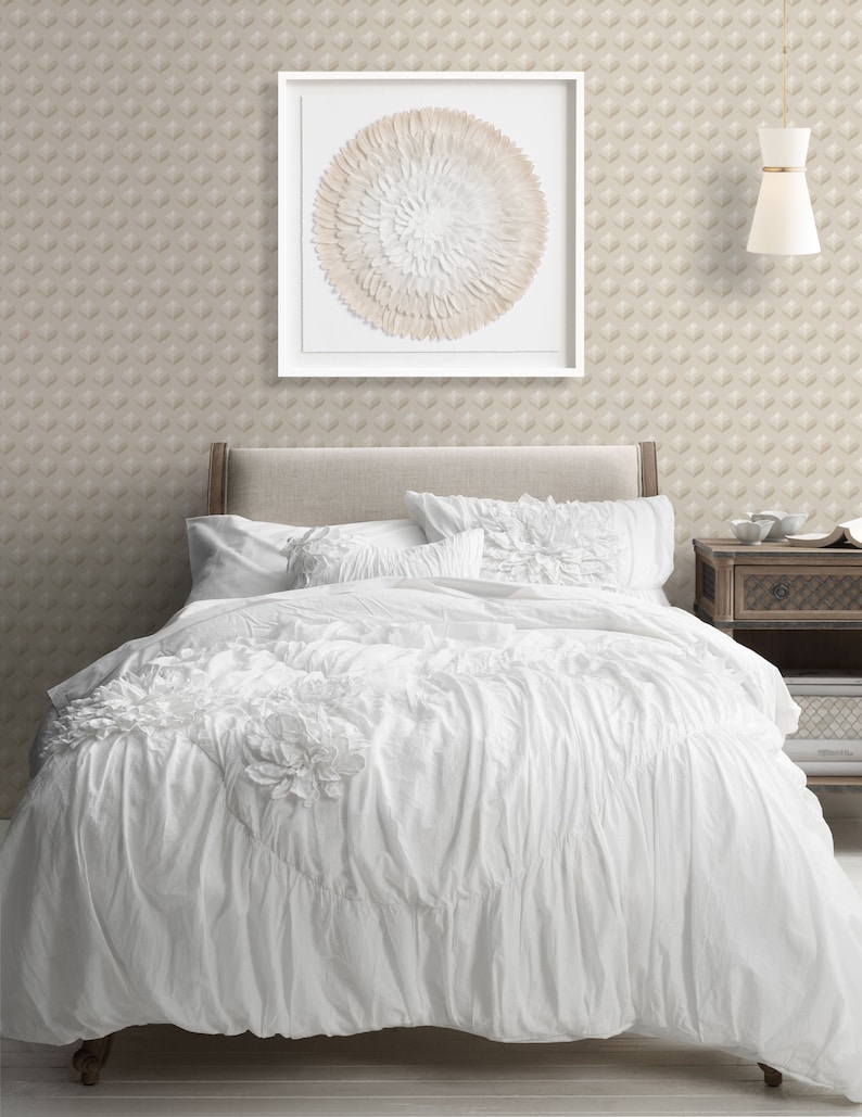 K&ouml;nnte beinhalten: Wei&szlig;er, ger&uuml;schter Bettbezug mit einem wei&szlig;en Kopfteil und zwei wei&szlig;en Kissen. &Uuml;ber dem Bett h&auml;ngt ein gerahmtes, rundes Wandbild mit einem wei&szlig;en und beigefarbenen Federdesign.