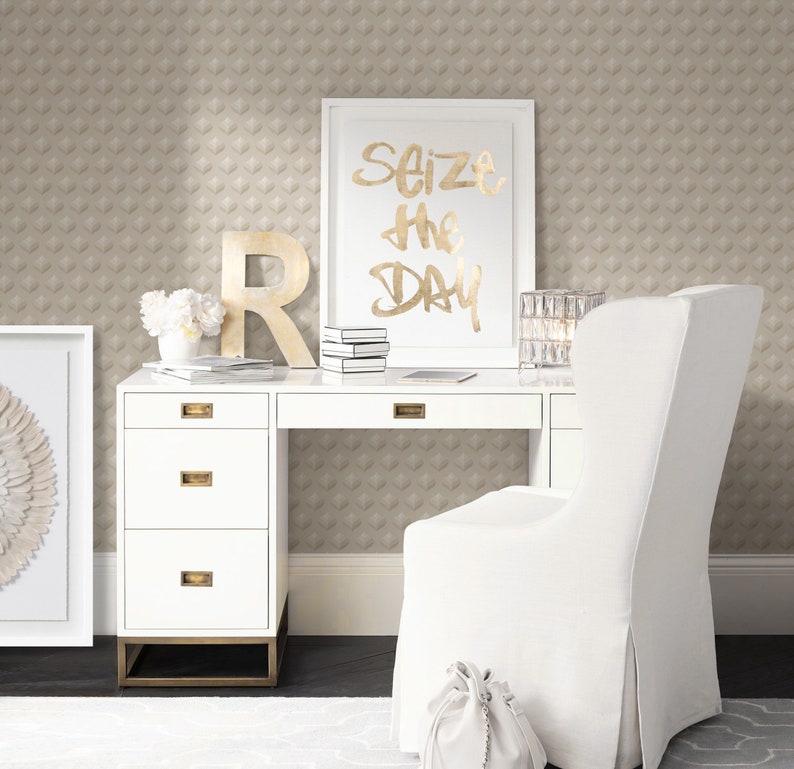 K&ouml;nnte beinhalten: Ein wei&szlig;er Schreibtisch mit vier Schubladen und einem wei&szlig;en Stuhl. Der Schreibtisch hat goldene Beschl&auml;ge und ein goldenes Gestell. Ein gerahmtes Bild mit dem Text "Seize the Day" h&auml;ngt an der Wand &uuml;ber dem Schreibtisch. Die Wand ist mit einer beigefarbenen Tapete mit geometrischem Muster bedeckt.