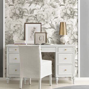 May include: A white desk with six drawers and gold knobs sits in front of a wall covered in a gray and white floral wallpaper. A white chair with a linen seat sits in front of the desk. A gold lamp with a white marble base sits on the desk.