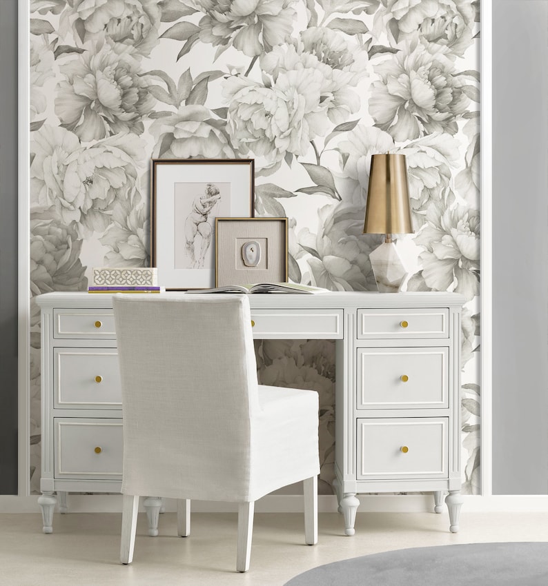 May include: A white desk with six drawers and gold knobs sits in front of a wall covered in a gray and white floral wallpaper. A white chair with a linen seat sits in front of the desk. A gold lamp with a white marble base sits on the desk.