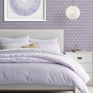K&ouml;nnte beinhalten: Ein Lavendelbett mit einem wei&szlig;en Kopfteil und einem wei&szlig;en und lavendelfarbenen, gemusterten Bettbezug. Das Bett ist mit einem wei&szlig;en Kissen und einem braunen herzf&ouml;rmigen Kissen bezogen. Ein wei&szlig; gerahmtes Bild mit einem wei&szlig;en und lavendelfarbenen Blumendesign h&auml;ngt an der Wand &uuml;ber dem Bett. Eine wei&szlig;e Lampe mit einem runden Schirm h&auml;ngt von der Decke. Ein wei&szlig;er Nachttisch mit einer Schublade steht rechts neben dem Bett.