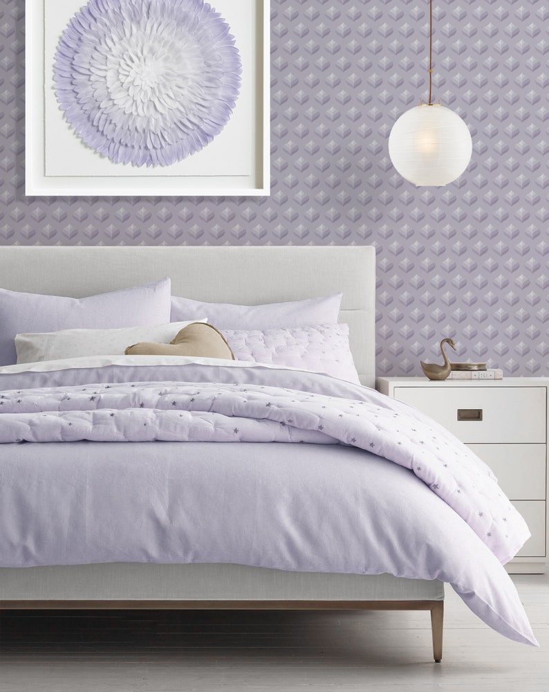 K&ouml;nnte beinhalten: Ein Lavendelbett mit einem wei&szlig;en Kopfteil und einem wei&szlig;en und lavendelfarbenen, gemusterten Bettbezug. Das Bett ist mit einem wei&szlig;en Kissen und einem braunen herzf&ouml;rmigen Kissen bezogen. Ein wei&szlig; gerahmtes Bild mit einem wei&szlig;en und lavendelfarbenen Blumendesign h&auml;ngt an der Wand &uuml;ber dem Bett. Eine wei&szlig;e Lampe mit einem runden Schirm h&auml;ngt von der Decke. Ein wei&szlig;er Nachttisch mit einer Schublade steht rechts neben dem Bett.