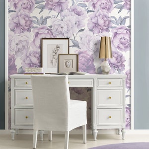 May include: A white desk with six drawers and gold knobs sits in front of a wall covered in a floral wallpaper featuring purple peonies. A white chair with a white cushion sits in front of the desk. A gold lamp with a white base sits on the desk.
