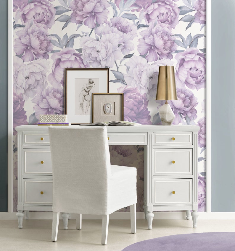 May include: A white desk with six drawers and gold knobs sits in front of a wall covered in a floral wallpaper featuring purple peonies. A white chair with a white cushion sits in front of the desk. A gold lamp with a white base sits on the desk.