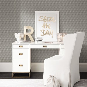 K&ouml;nnte beinhalten: Ein wei&szlig;er Schreibtisch mit vier Schubladen und goldenen Beschl&auml;gen. Ein wei&szlig;er Stuhl mit einem wei&szlig;en Stoffbezug steht vor dem Schreibtisch. Ein gerahmter Druck mit dem Text "Seize the Day" h&auml;ngt an der Wand hinter dem Schreibtisch. Die Wand ist mit einer grauen Tapete mit geometrischem Muster bedeckt.