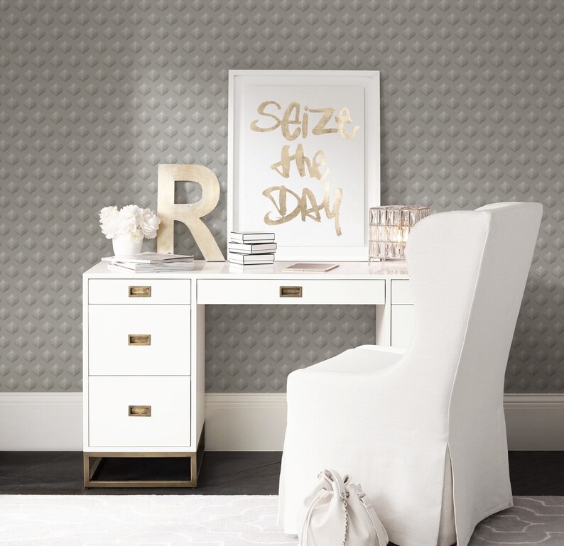 K&ouml;nnte beinhalten: Ein wei&szlig;er Schreibtisch mit vier Schubladen und goldenen Beschl&auml;gen. Ein wei&szlig;er Stuhl mit einem wei&szlig;en Stoffbezug steht vor dem Schreibtisch. Ein gerahmter Druck mit dem Text "Seize the Day" h&auml;ngt an der Wand hinter dem Schreibtisch. Die Wand ist mit einer grauen Tapete mit geometrischem Muster bedeckt.