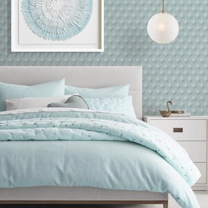 K&ouml;nnte beinhalten: Ein hellblaues Bett mit einem wei&szlig;en Kopfteil und einem hellblauen und wei&szlig;en gemusterten Bettbezug. Das Bett steht in einem Schlafzimmer mit einer hellblauen und wei&szlig;en gemusterten Tapete und einem wei&szlig;en gerahmten Bild mit einem hellblauen und wei&szlig;en gemusterten Kreis in der Mitte. Ein wei&szlig;er Nachttisch mit einer Schublade befindet sich rechts neben dem Bett. Eine wei&szlig;e Globus-Pendelleuchte h&auml;ngt von der Decke.