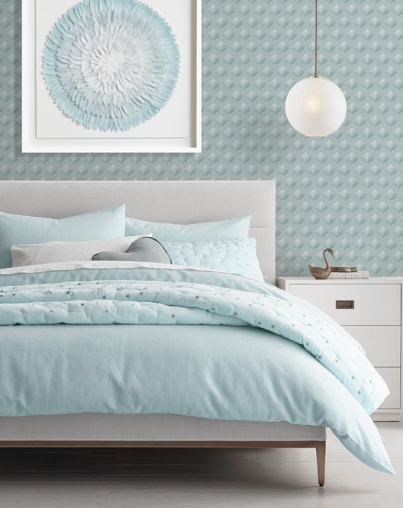 K&ouml;nnte beinhalten: Ein hellblaues Bett mit einem wei&szlig;en Kopfteil und einem hellblauen und wei&szlig;en gemusterten Bettbezug. Das Bett steht in einem Schlafzimmer mit einer hellblauen und wei&szlig;en gemusterten Tapete und einem wei&szlig;en gerahmten Bild mit einem hellblauen und wei&szlig;en gemusterten Kreis in der Mitte. Ein wei&szlig;er Nachttisch mit einer Schublade befindet sich rechts neben dem Bett. Eine wei&szlig;e Globus-Pendelleuchte h&auml;ngt von der Decke.
