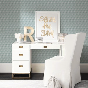 K&ouml;nnte beinhalten: Wei&szlig;er Schreibtisch mit vier Schubladen und goldenen Beschl&auml;gen. Ein wei&szlig;er Stuhl mit einer wei&szlig;en Tasche auf der Sitzfl&auml;che. Ein gerahmter Druck mit dem Text "Seize the Day" in goldenen Buchstaben. Die Wand hinter dem Schreibtisch ist mit einer hellblauen Tapete mit geometrischem Muster bedeckt.