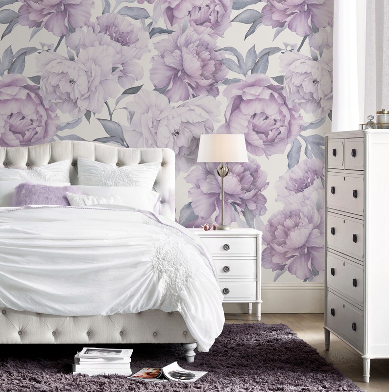 May include: A white upholstered bed with a tufted headboard and a white nightstand with three drawers. The bed is dressed in white bedding and a lavender throw pillow. The wall behind the bed is covered in a floral wallpaper with large purple peonies.