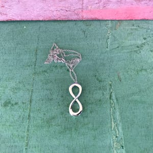 May include: A silver-colored infinity symbol pendant necklace. The pendant is a polished, smooth figure eight shape. The necklace chain is delicate and silver. The background is a green wooden surface with a pink wooden edge.