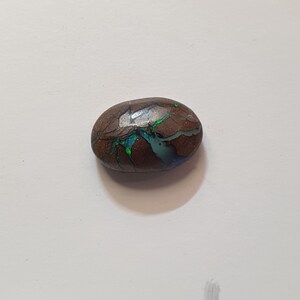 Könnte beinhalten: Ein polierter, oval geformter Edelstein mit brauner Grundfarbe. Der Stein weist komplizierte Muster aus grünen, blauen und irisierenden Farben auf, die ein einzigartiges und auffälliges Aussehen erzeugen. Es handelt sich wahrscheinlich um einen Opal.