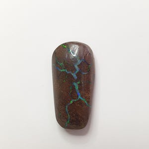 May include: A polished, oblong gemstone with a dark brown base color. The stone features a striking pattern of iridescent blue and green veins, creating a unique, natural design. The gemstone is set against a plain white background.