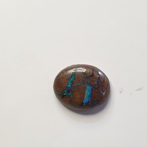 May include: A brown, oval-shaped gemstone with a cracked pattern. The cracks are filled with iridescent blue and green hues, creating a striking contrast against the dark brown base. The gemstone is set against a white background.