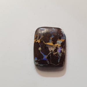 May include: A rectangular, polished gemstone with a dark brown base color. The stone features intricate patterns of yellow, blue, and white, resembling a natural landscape. The surface is glossy, highlighting the unique internal structures and colors.