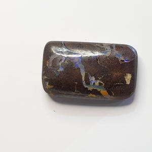 May include: A rectangular, polished opal stone with a dark brown base color. The stone features iridescent blue, yellow, and white patterns across its surface. The opal has rounded edges and a smooth, glossy finish.