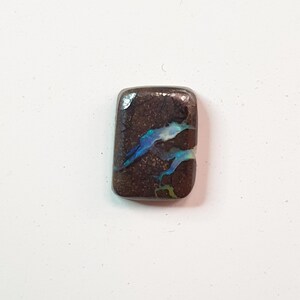 May include: A rectangular, polished gemstone with a dark brown base color. The stone features iridescent blue and green patterns, creating a striking visual contrast. The gemstone is likely used in jewelry or as a decorative element.