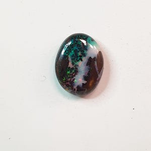 Könnte beinhalten: Ein ovales Schmuckstück mit dunkler Grundfarbe, das irisierende grüne, blaue und violette Muster aufweist. Die polierte Oberfläche reflektiert das Licht und zeigt ein Farbspiel. Der Edelstein liegt vor einem weißen Hintergrund.