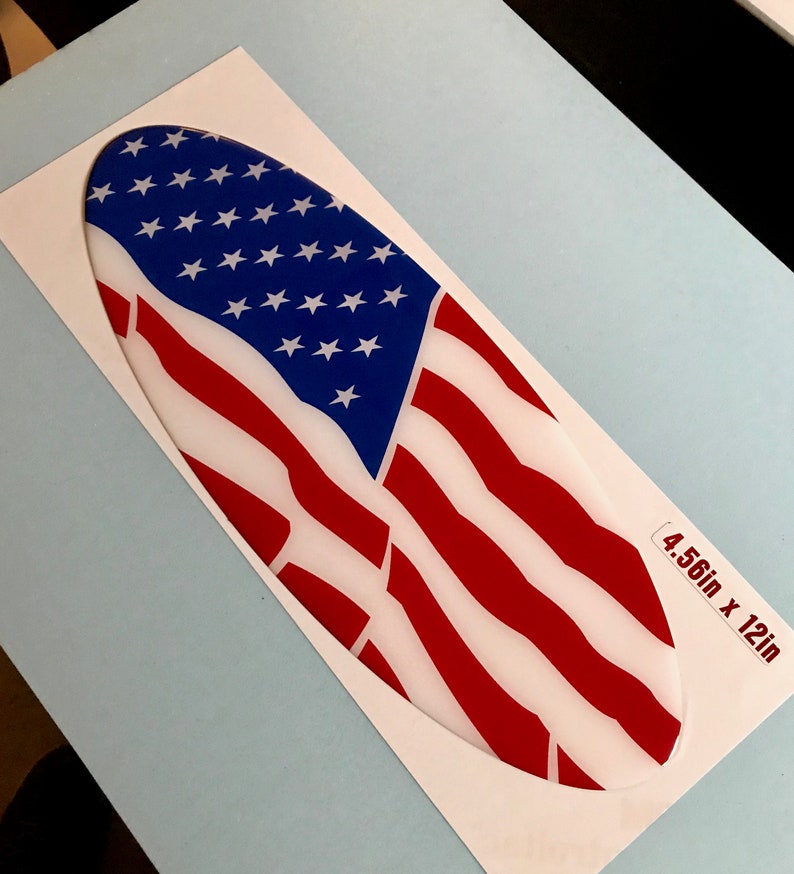 May include: Oval-shaped sticker with an American flag design. The flag is red, white, and blue with a white background. The sticker is 4.56 inches by 12 inches.