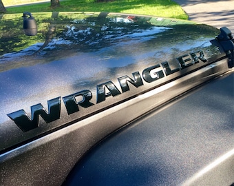 Könnte beinhalten: Nahaufnahme eines schwarzen "WRANGLER"-Aufklebers auf einer dunkelgrauen Motorhaube.