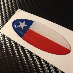 May include: A white oval sticker with a 3D raised design of the Texas flag. The flag is blue, white, and red with a white star in the blue section.