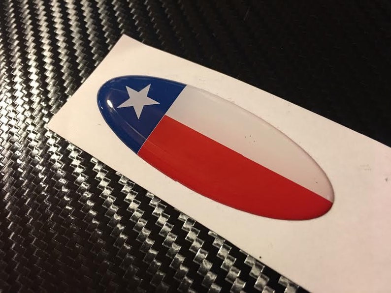May include: A white oval sticker with a 3D raised design of the Texas flag. The flag is blue, white, and red with a white star in the blue section.