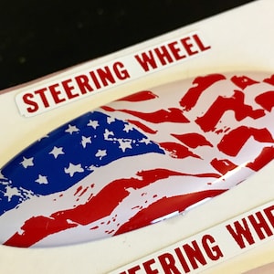 May include: A white oval-shaped sticker with a distressed American flag design in red, white, and blue. The sticker has the words "Steering Wheel" printed on either side.