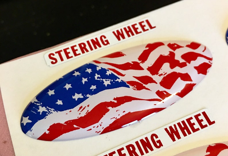May include: A white oval-shaped sticker with a distressed American flag design in red, white, and blue. The sticker has the words "Steering Wheel" printed on either side.
