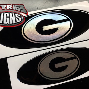 May include: Two black oval-shaped decals with a silver metallic 'G' inside a white circle. The decals are on white paper.