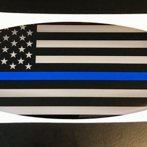 May include: Oval-shaped car decal with a black and white American flag design with a thin blue line across the middle. The decal measures 3.25 inches by 8.25 inches.
