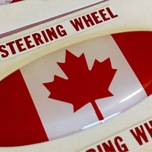 May include: A red, white, and red Canadian flag sticker with a white oval shape and the words "Steering Wheel" on a white background.