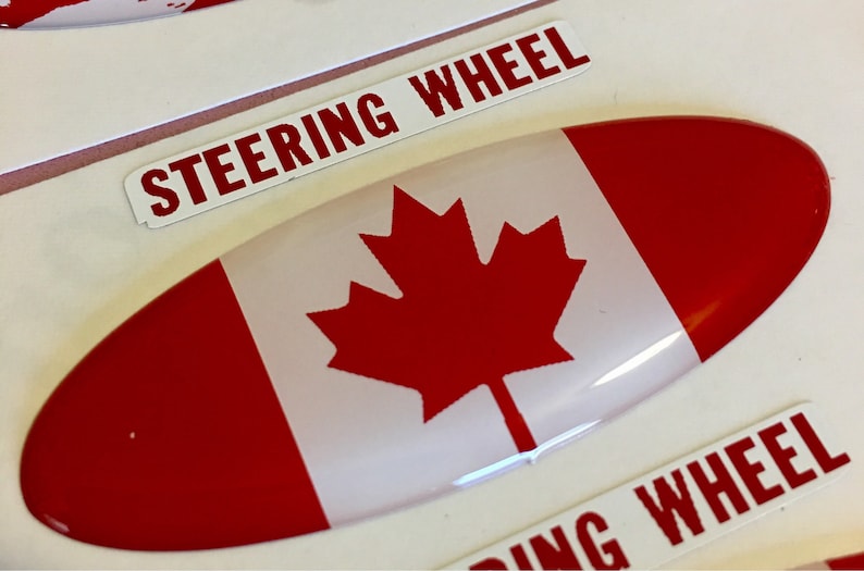 May include: A red, white, and red Canadian flag sticker with a white oval shape and the words "Steering Wheel" on a white background.