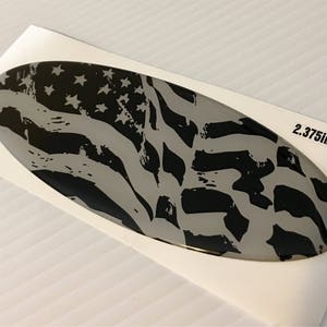 May include: A black and white distressed American flag design on a silver oval sticker. The sticker measures 2.375 inches by 6.3 inches.