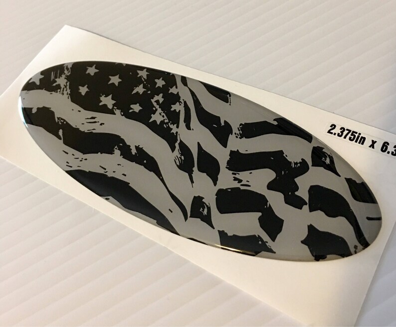 May include: A black and white distressed American flag design on a silver oval sticker. The sticker measures 2.375 inches by 6.3 inches.