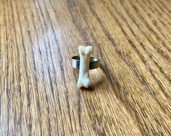 May include: A unique ring featuring a small, off-white bone set on a silver-colored band. The bone is vertically oriented, showcasing its natural shape and texture. The ring is displayed on a wooden surface, highlighting the contrast between the organic and metallic elements.