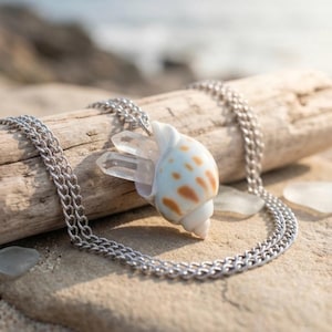 May include: A necklace featuring a white seashell pendant with brown spots and a clear, pointed quartz crystal protruding from the opening. The pendant is attached to a thin, silver-toned metal chain with small, interlocking oval links.