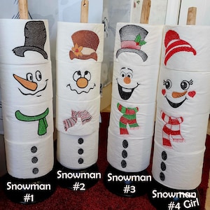 May include: Four white toilet paper rolls decorated with embroidered snowman designs. Each roll has a different snowman design with a hat, scarf, and facial features. The designs are numbered 1 through 4. The fourth snowman is labeled "Girl".