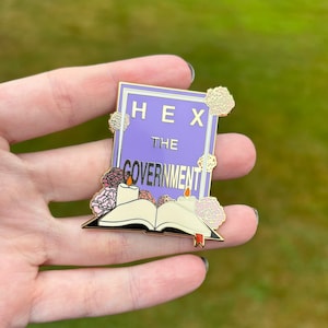 May include: A gold-colored enamel pin with a purple background and the text "HEX THE GOVERNMENT". The pin features a book with two candles and flowers.