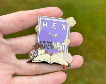 May include: A gold-colored enamel pin with a purple background and the text "HEX THE GOVERNMENT". The pin features a book with two candles and flowers.