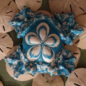 Puede incluir: Una figurita de rana de cerámica azul con un diseño de flor blanca. La rana está rodeada de dólares de arena.