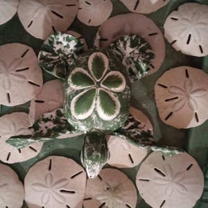 May include: A green and white painted turtle figurine surrounded by a collection of sand dollar shells. The turtle has a five-petal flower design on its shell.