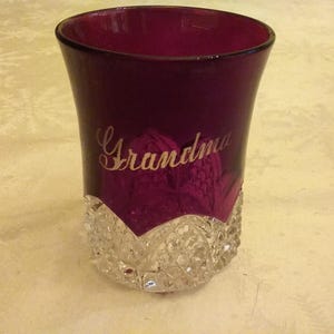 May include: A burgundy-colored glass with a clear, cut-glass base. The word "Grandma" is etched in gold script on the glass. The glass has a flared shape and is likely a decorative or collectible item.