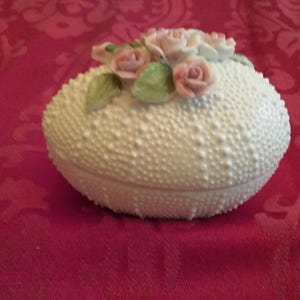 May include: A white, textured ceramic trinket box with a lid, adorned with pink rose and green leaf accents. The box has a rounded shape and sits on a red patterned surface. A decorative item.