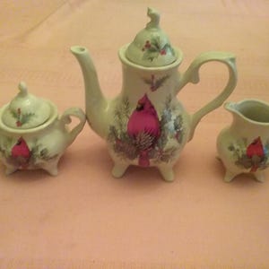 May include: A three-piece ceramic tea set with a cream-colored finish. The set includes a teapot, a sugar bowl with a lid, and a creamer. Each piece features a vibrant red cardinal bird design, surrounded by winter foliage.