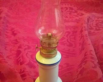 May include: A white ceramic oil lamp with a blue trim and a clear glass chimney. The lamp has a gold-colored metal wick holder and a small handle on the side.