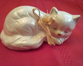 May include: A white porcelain figurine of a sleeping cat with a cream-colored satin ribbon tied around its neck.